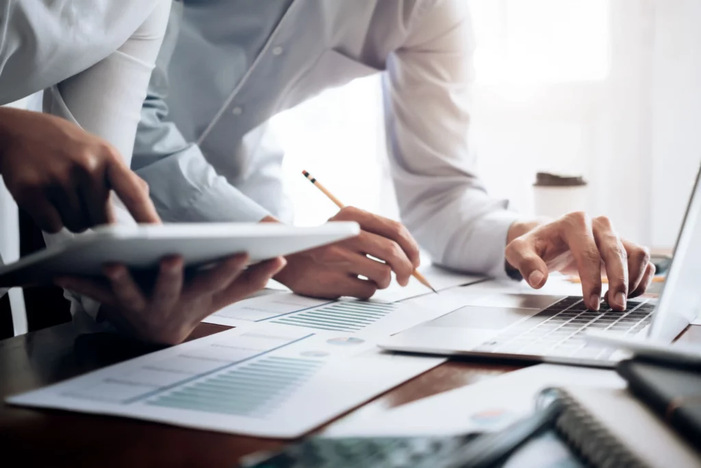 Zwei Personen arbeiten an Finanzdokumenten mit Laptop und Stift.